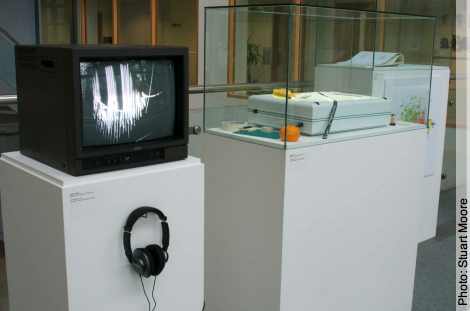 Photo with box monitor showing film still, on plinth with headphones; to right: glass cabinet with lightbox on plinth 